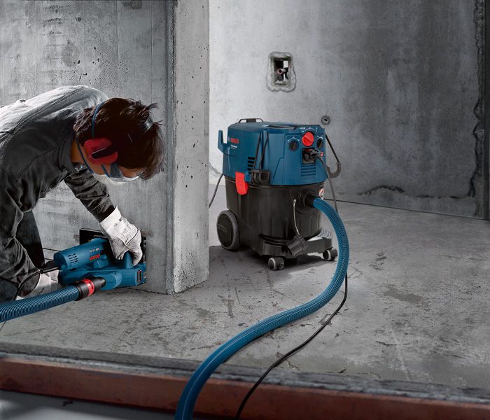 Eine Person in Schutzausrüstung schleift Beton mit einem Schlauchanschluss-Staubabsauggerät., Industriesauger, Baustellensauger, blau-schwarz, Saugschlauch, Werkzeugabsaugung