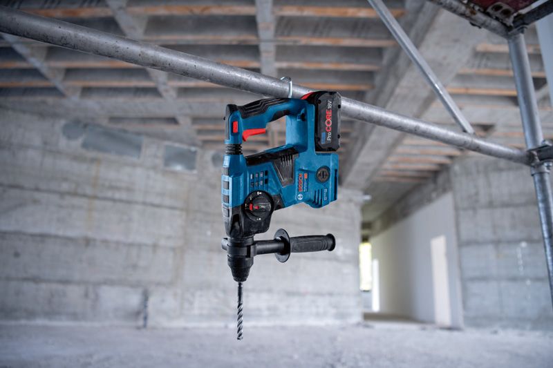 Akku-Bohrhammer, der am Griff an einem Metallgerüst in einem Betongebäude hängt., Akku-Bohrhammer, Bohrmaschine, blau-schwarz, Zusatzhandgriff, SDS-Bohrer