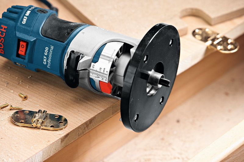 Handfräse in der Nähe von Türscharnieren und Schrauben auf einer Holzoberfläche positioniert., Kantenfräse, Elektrowerkzeug, blau-schwarz, Kunststoffgehäuse, Frästeller